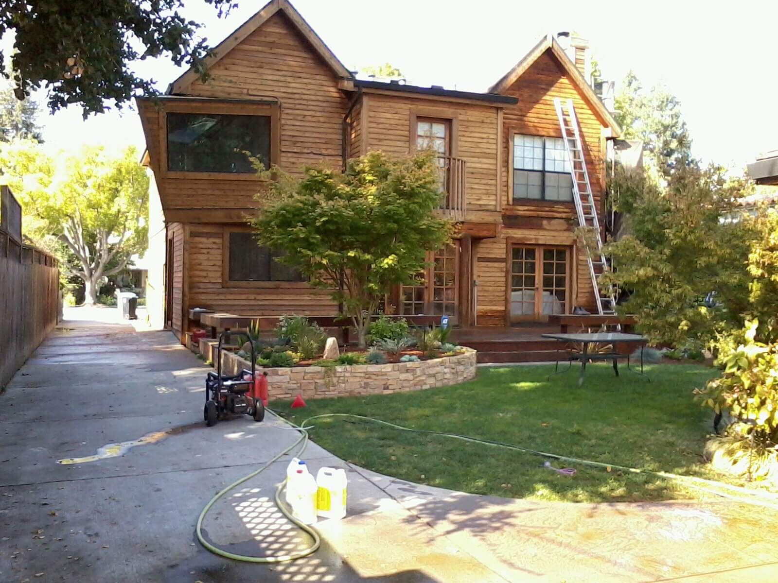Two-story wood-sided home mid-project with ladder and pressure washer during exterior surface prep
