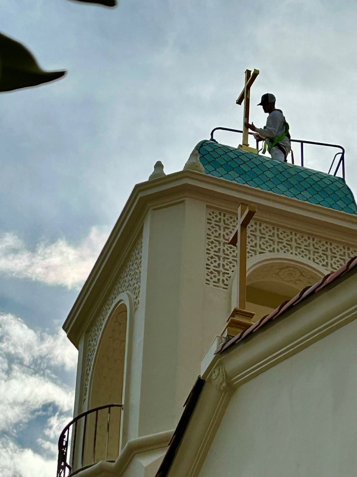 Craftsman in safety harness applying 24k gold leaf to the cross atop the teal-tiled cupola of St. Nicholas Church in Los Altos